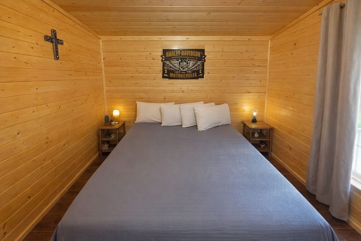 A wooden-paneled room with a bed, two bedside tables with lamps, a cross on the wall, and a Harley-Davidson motorcycle emblem.