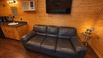 A wooden cabin interior with a leather sofa, a sink, a stove, and a television mounted on the wall.