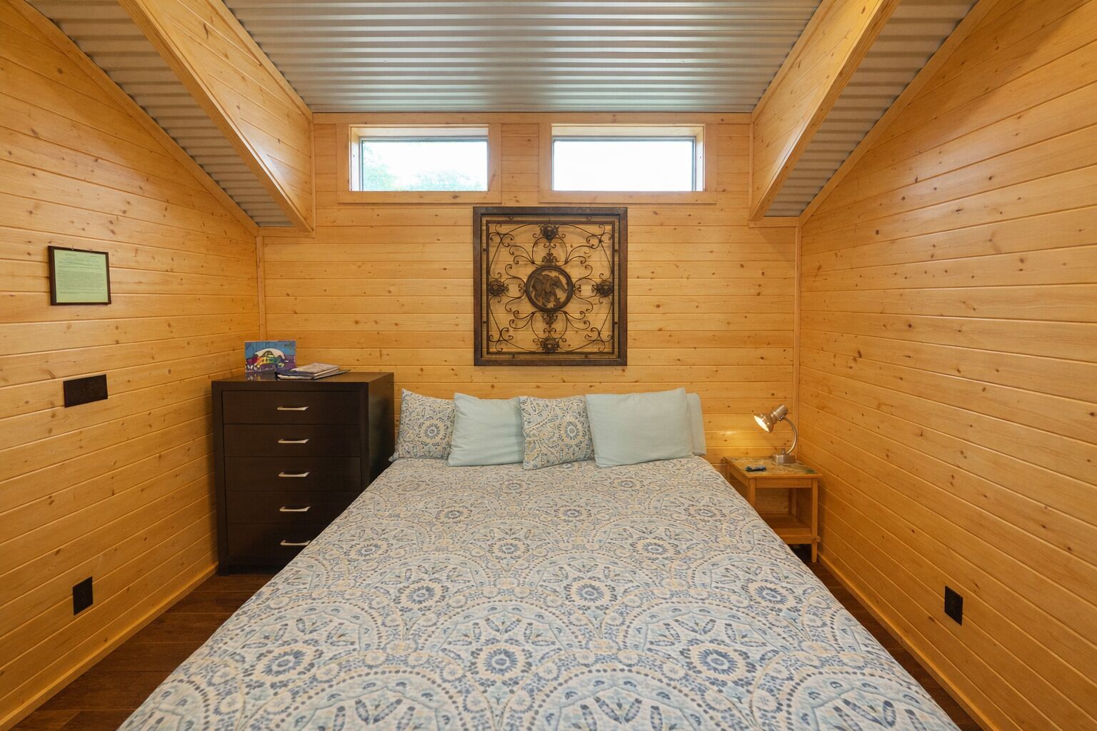 Camera da letto in una baita di legno, con un letto, una cassettiera, due finestre e un'opera d'arte appesa al muro.