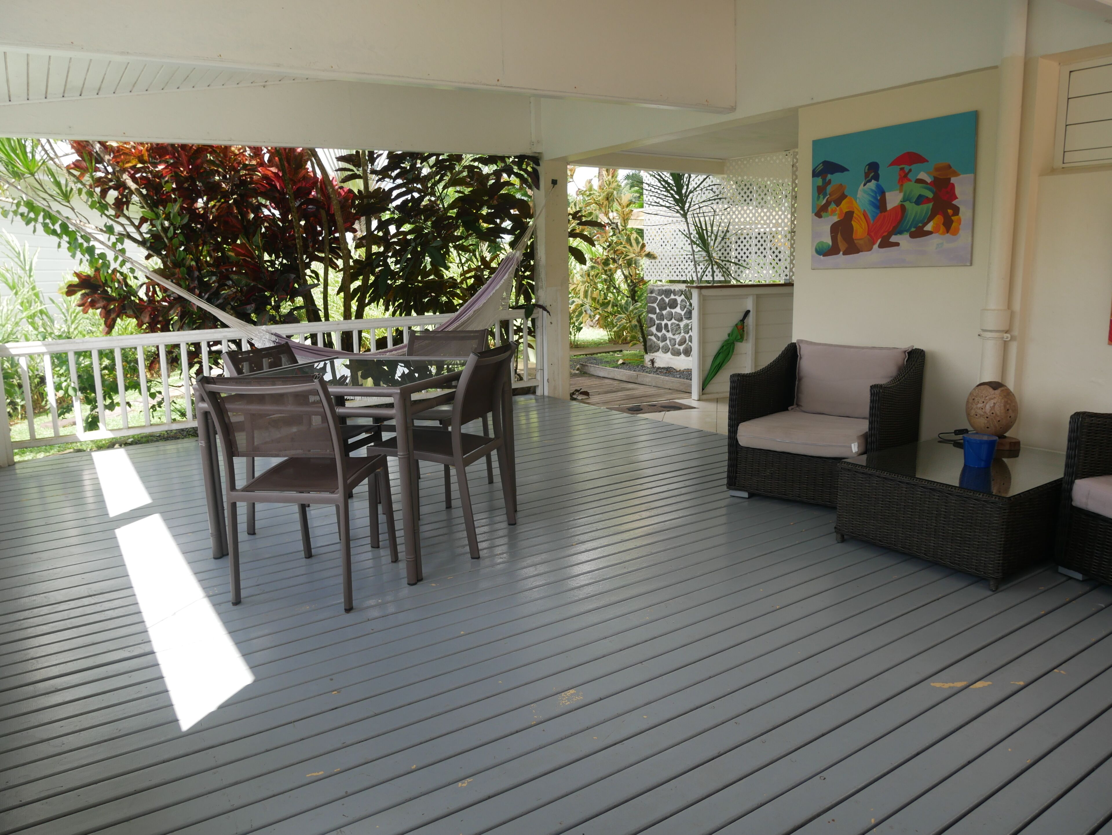 A covered outdoor area with a table and chairs, a wicker sofa, and a painting on the wall.