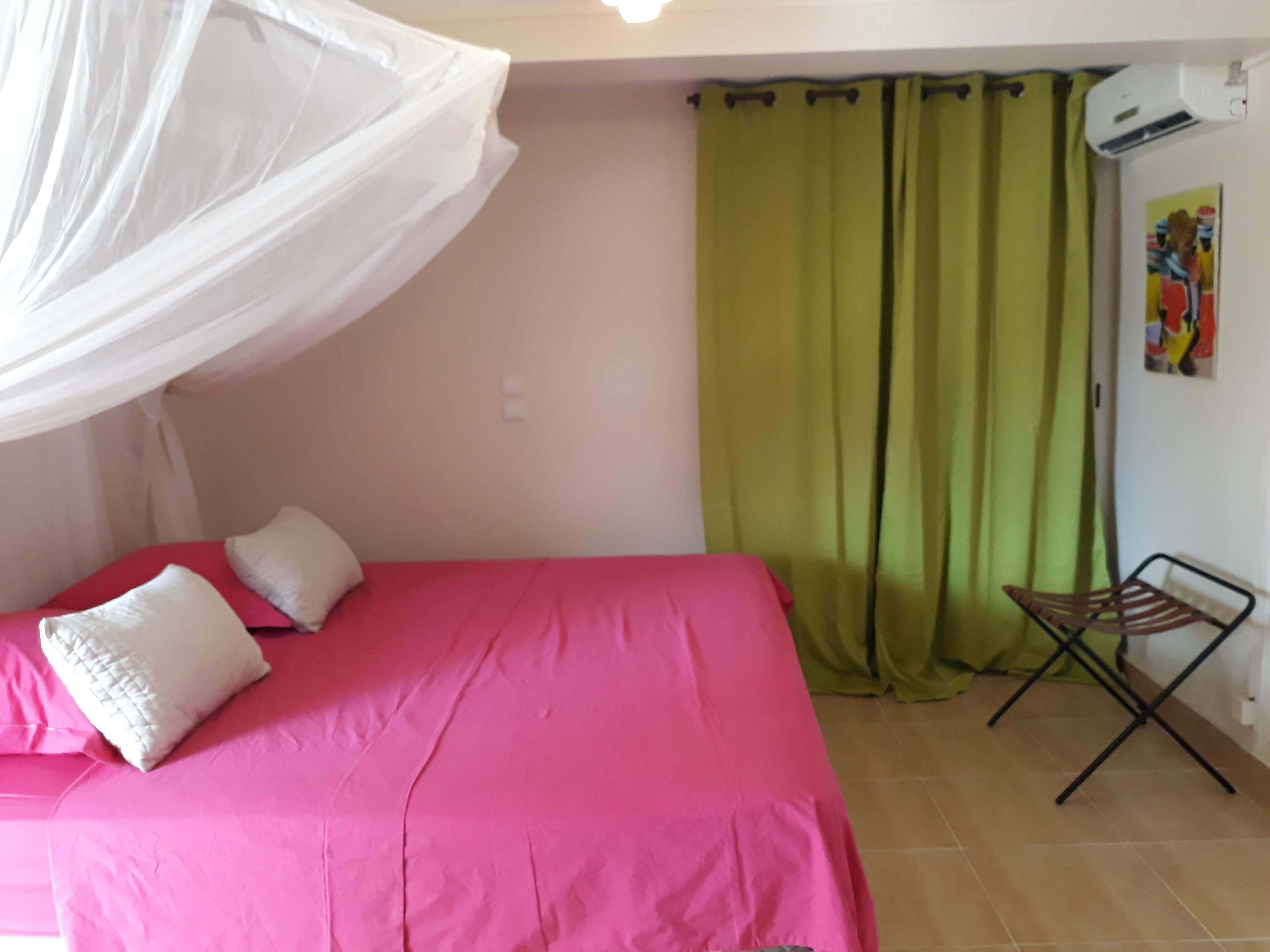 A bedroom with a bed covered in pink bedding, white pillows, and a white canopy. There is a green curtain and a small folding chair in the corner.
