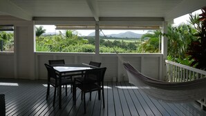 A covered outdoor area with a table and chairs, a hammock, and a view of a lush landscape with mountains in the distance. - CITRONELLA APARTMENT (PETIT-BOURG)
