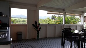 A room with a wooden floor, a dining table with chairs, a small trash bin, and a window with a view of greenery and mountains. - CITRONELLA APARTMENT (PETIT-BOURG)