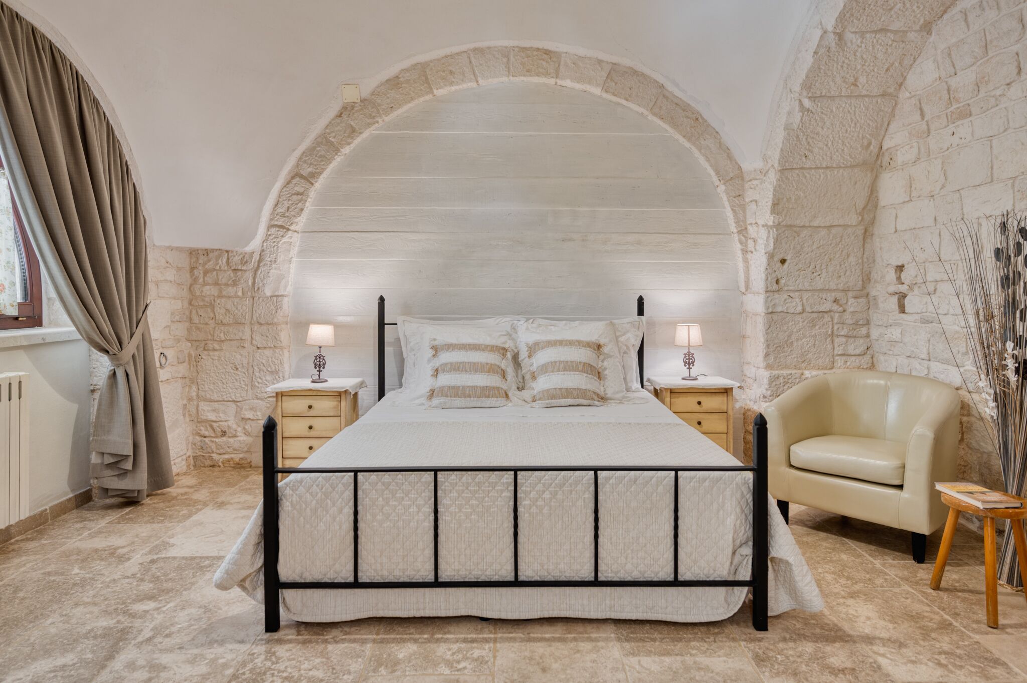 A bedroom with a stone archway, a bed with a quilted headboard, two bedside tables with lamps, a beige armchair, and a small wooden side table.
