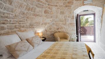 A bedroom with a stone wall, a bed with patterned bedding, a chair, and a nightstand with a lamp.