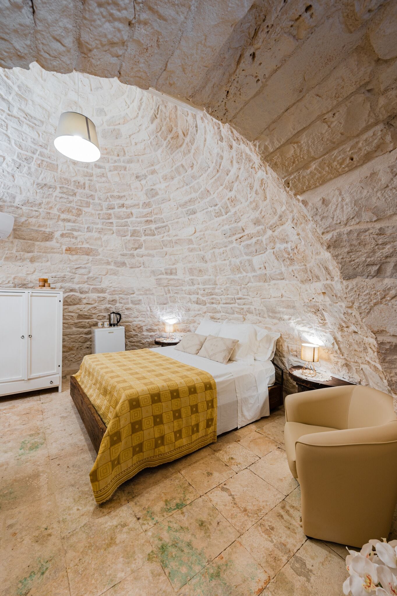 A bedroom with a stone ceiling, a bed with a yellow blanket, a white wardrobe, and a nightstand with a lamp.