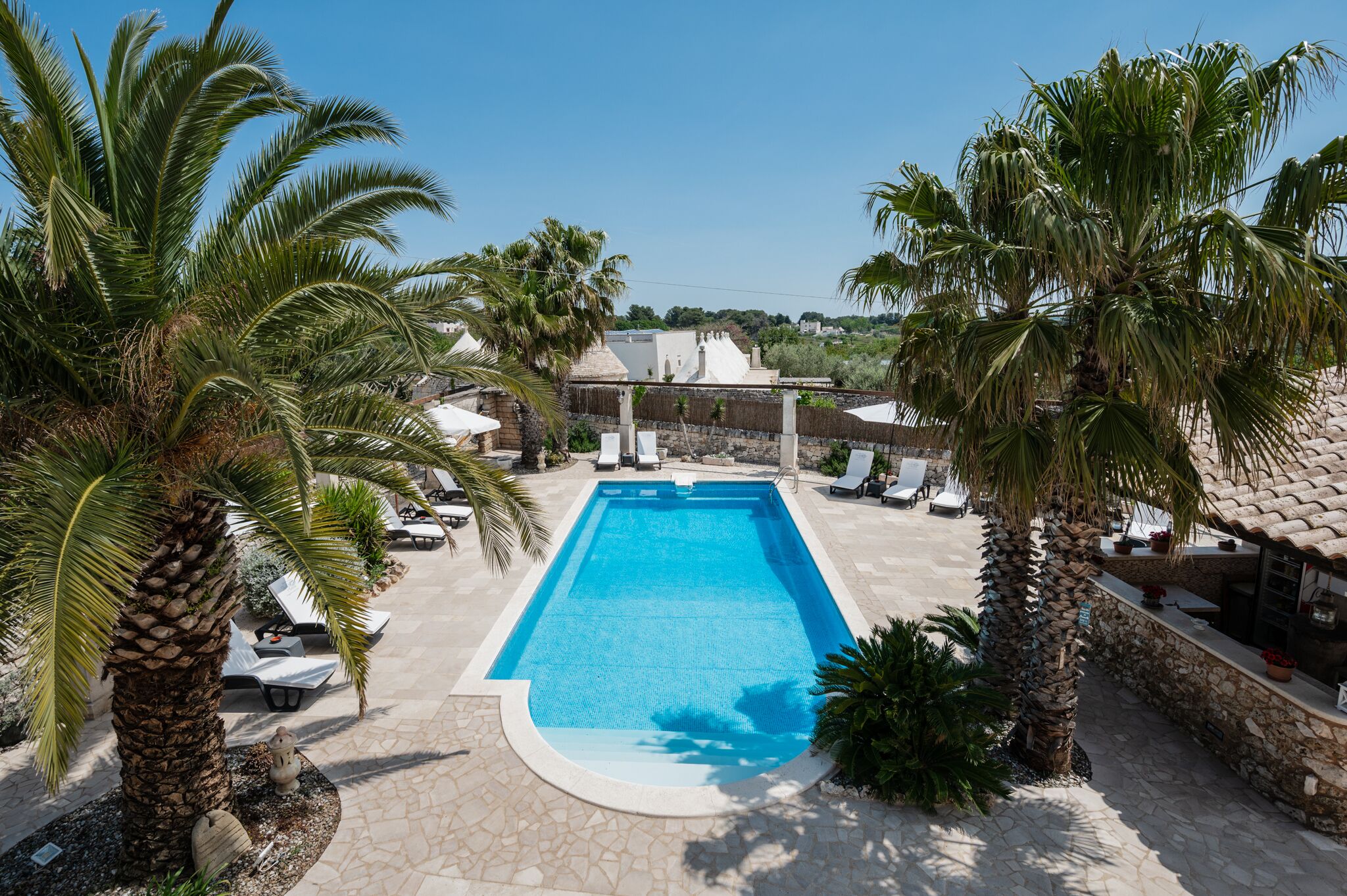 A rectangular swimming pool surrounded by lounge chairs and palm trees, with a stone patio and a building in the background.