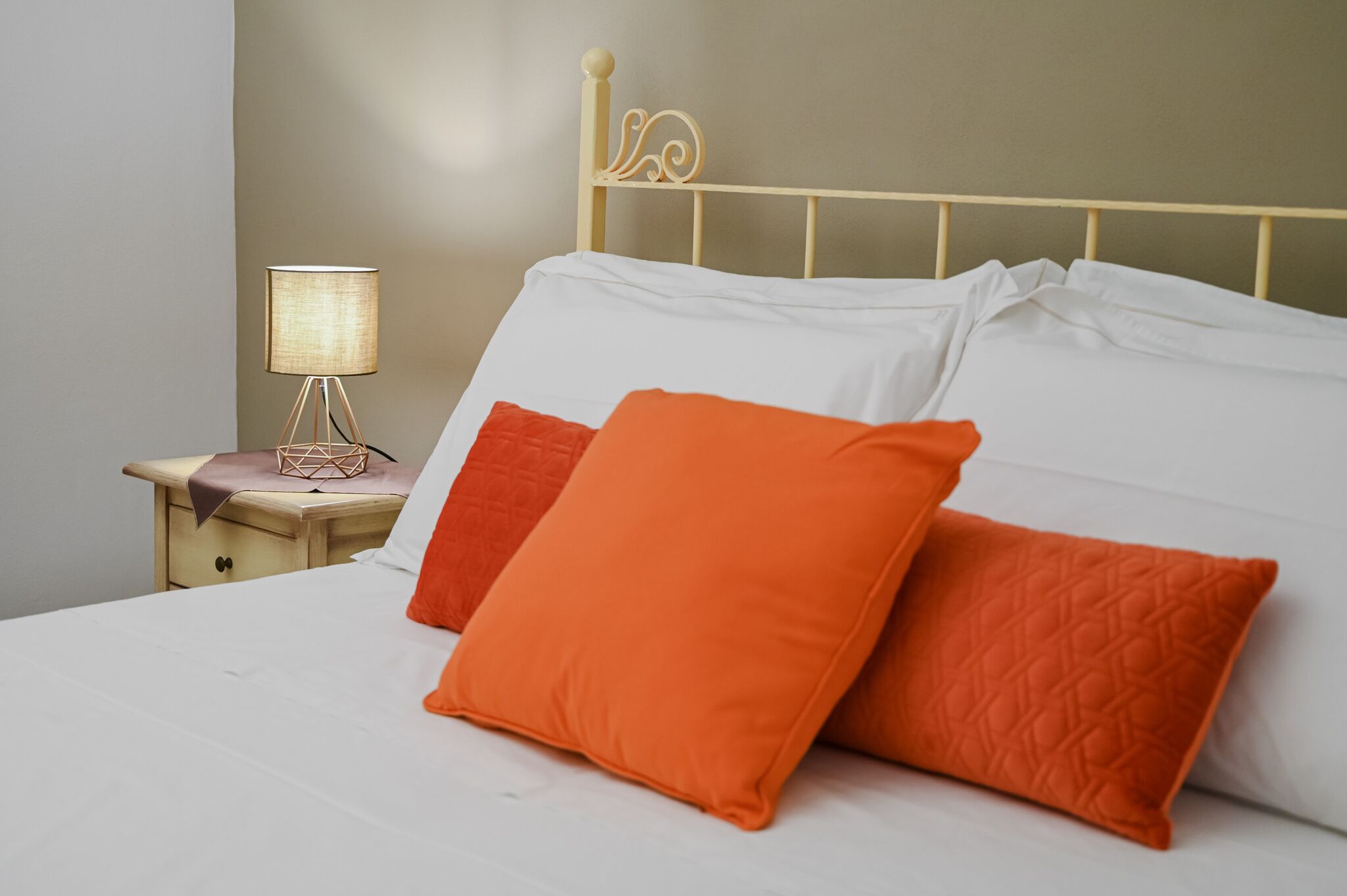 A neatly made bed with white and orange pillows, a bedside lamp, and a wooden nightstand.