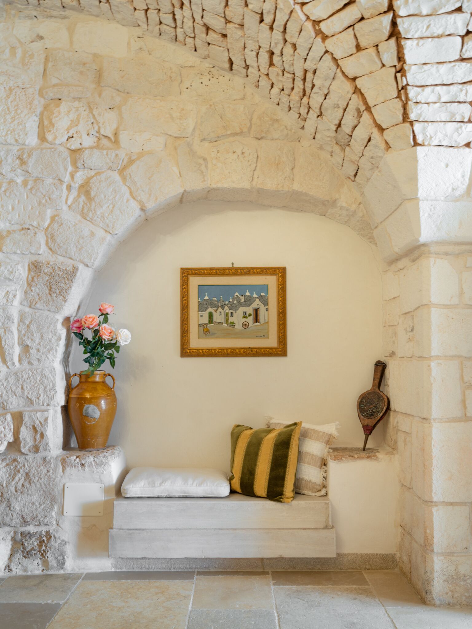 A stone-walled alcove with a cushioned bench, a framed painting, a vase with flowers, and a traditional wind chime.