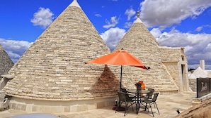 A stone-roofed building with a round table and chairs under an orange umbrella. - Trulli D'Autore (Martina Franca)