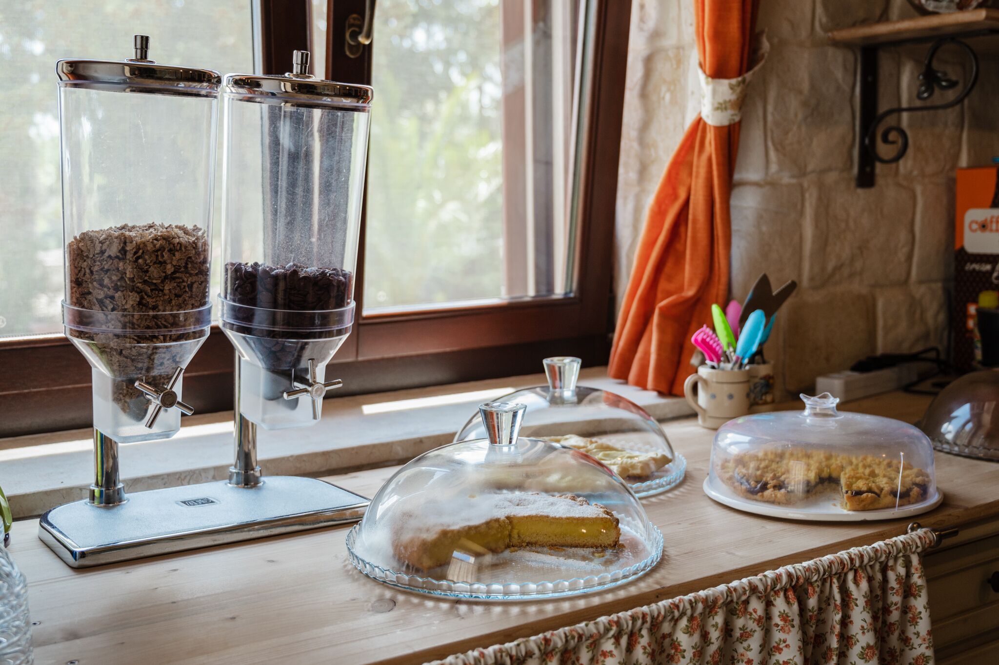 A buffet setup with two coffee dispensers, a pie covered with a glass dome, and a coffee mug on a table near a window.