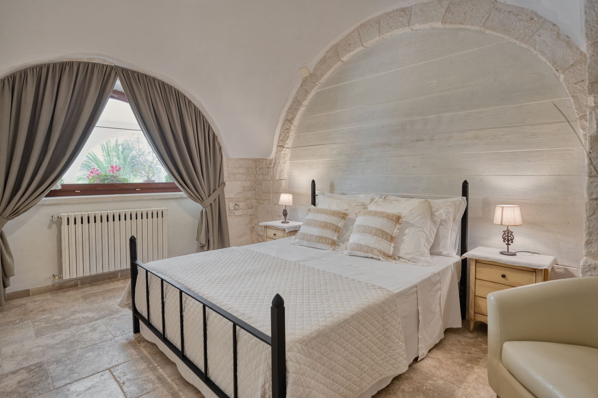 Een slaapkamer met een bed, twee nachtkastjes met lampen, een raam met gordijnen en een gewelfde stenen muur.