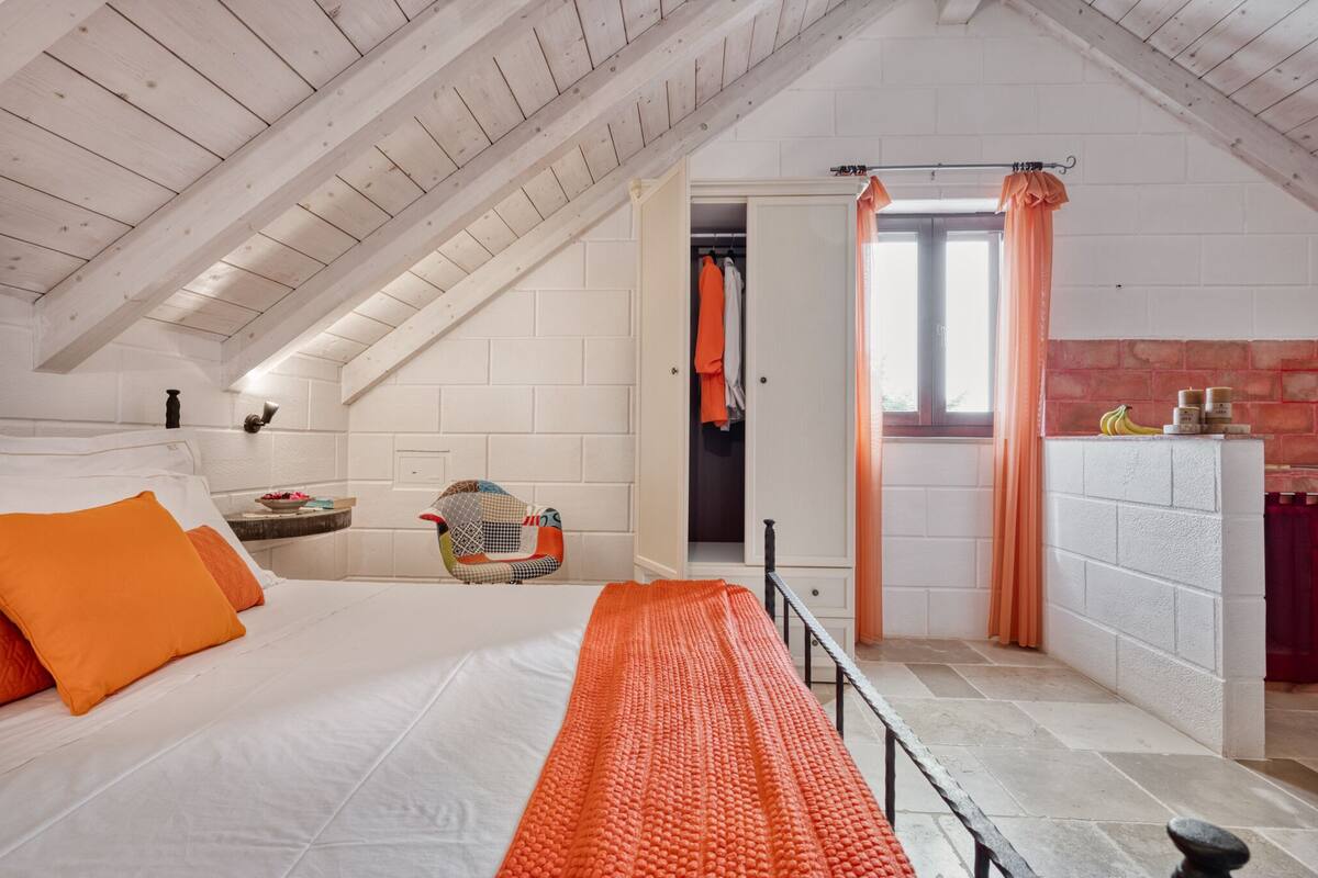 Et soveværelse med skråvæg, seng med orange sengetøj, en stol, et lille bord og et vindue med gardiner.