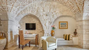 A stone-walled room with a television, a wicker chair, a sofa, and a table with a vase of flowers. - Trulli D'Autore (Martina Franca)