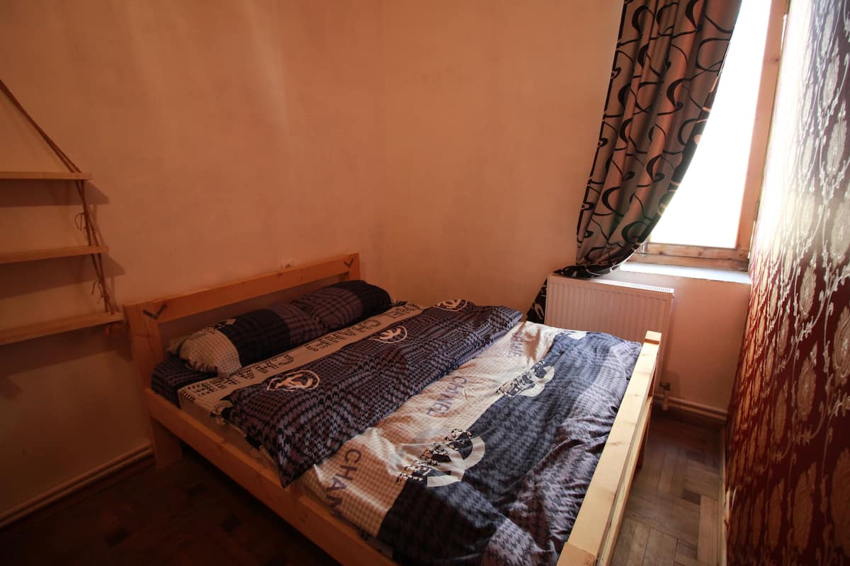 Un lit simple avec une couvre-lit à motifs, une tête de lit en bois et une fenêtre ornée de rideaux à motifs.