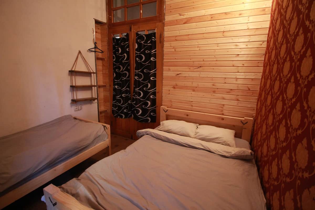 Une petite pièce chaleureuse avec deux lits simples, des murs en bois, un luminaire suspendu et un rideau à motifs.