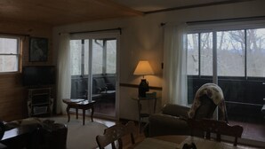 Ein gemütliches Wohnzimmer mit Holzbalkendecke, Fernseher, Lampe und großen Fenstern mit Vorhängen.