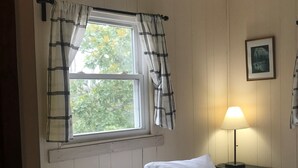 Ein ordentlich gemachtes Bett mit weißen Leinen in einem Zimmer mit Holzvertäfelung, einem Fenster mit Karottenvorhängen und einer Nachttischlampe.