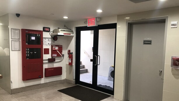 Un panel de control de alarma contra incendios, un extintor y una puerta de almacenamiento en un pasillo.