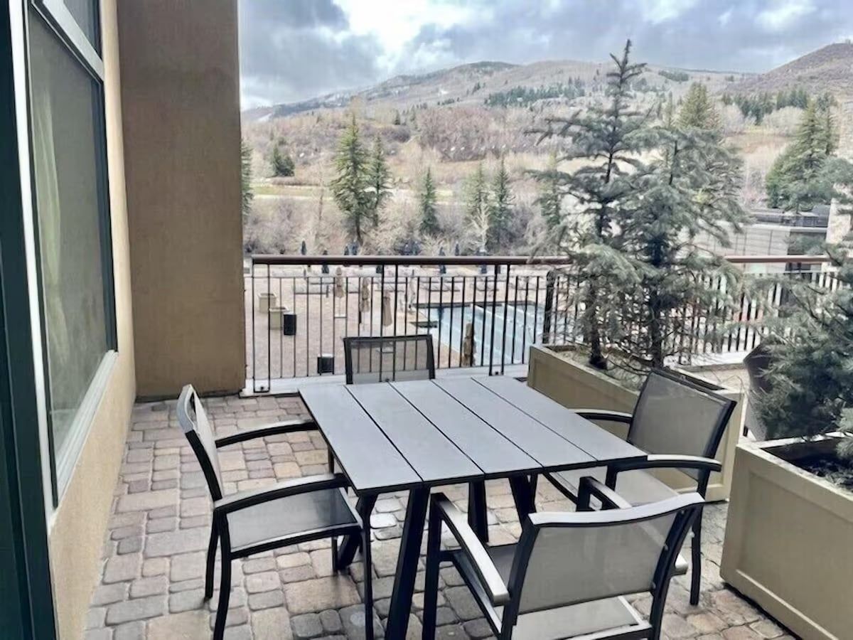 A balcony with a table and chairs, a view of trees and hills, and a metal railing.