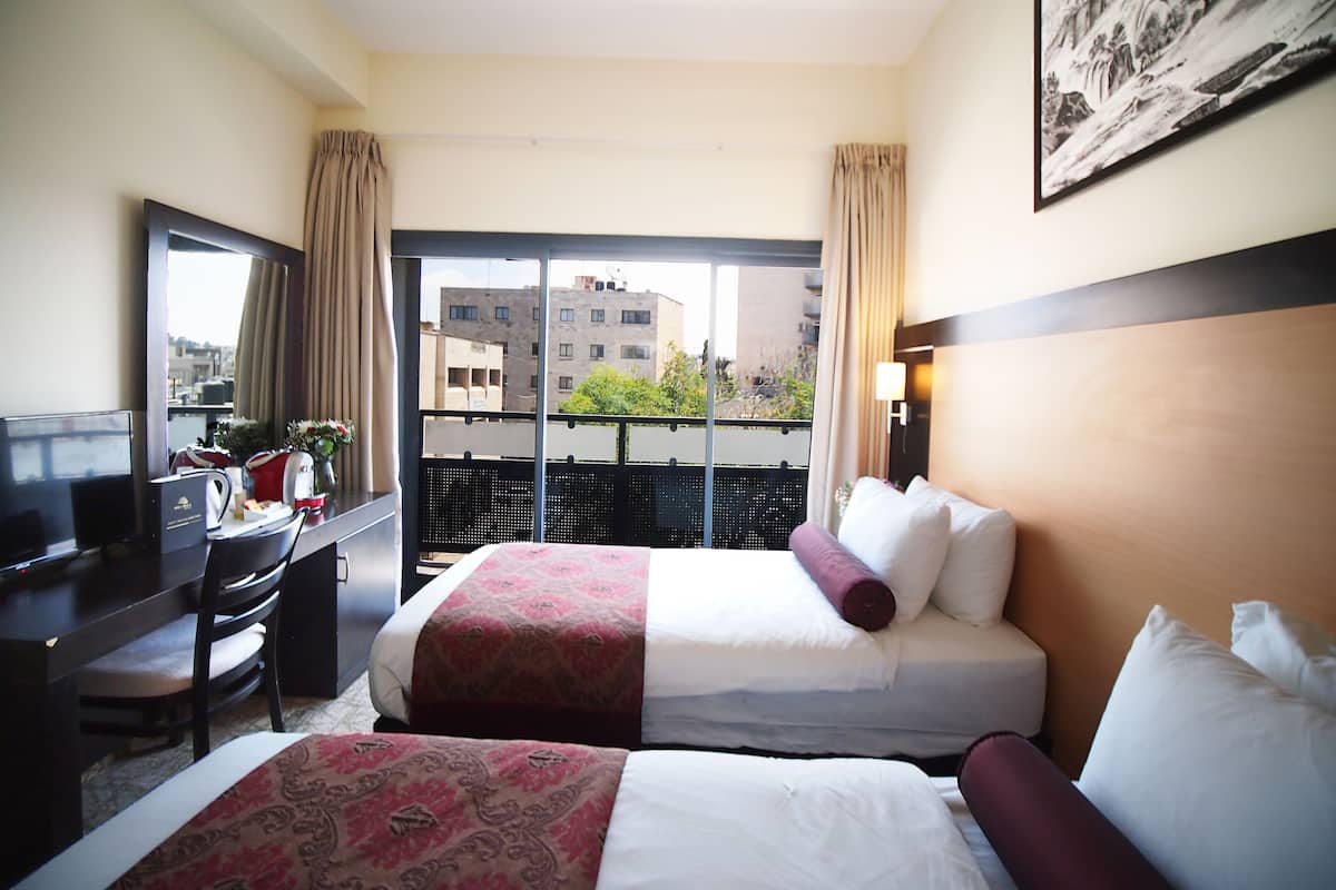A hotel room with two beds, a desk, a chair, and a large window with a view of buildings.
