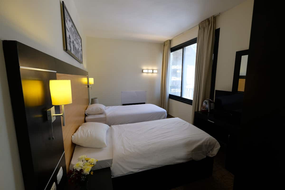 A hotel room with two single beds, a desk with a lamp, a TV, and a window with curtains.