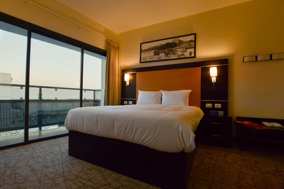 A hotel room with a large bed, a nightstand, a lamp, and a window with a view of the city.