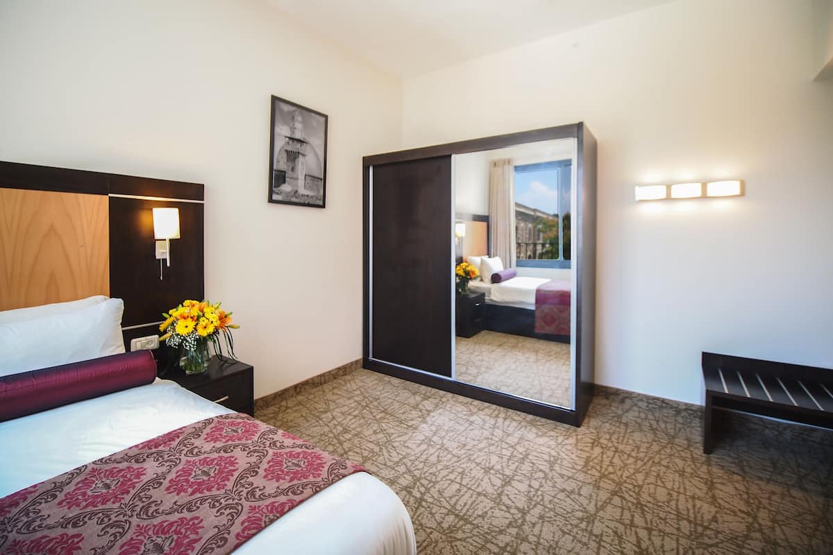 A hotel room with a bed, a nightstand with a vase of flowers, a desk with a lamp, a mirror, and a view of the outside.