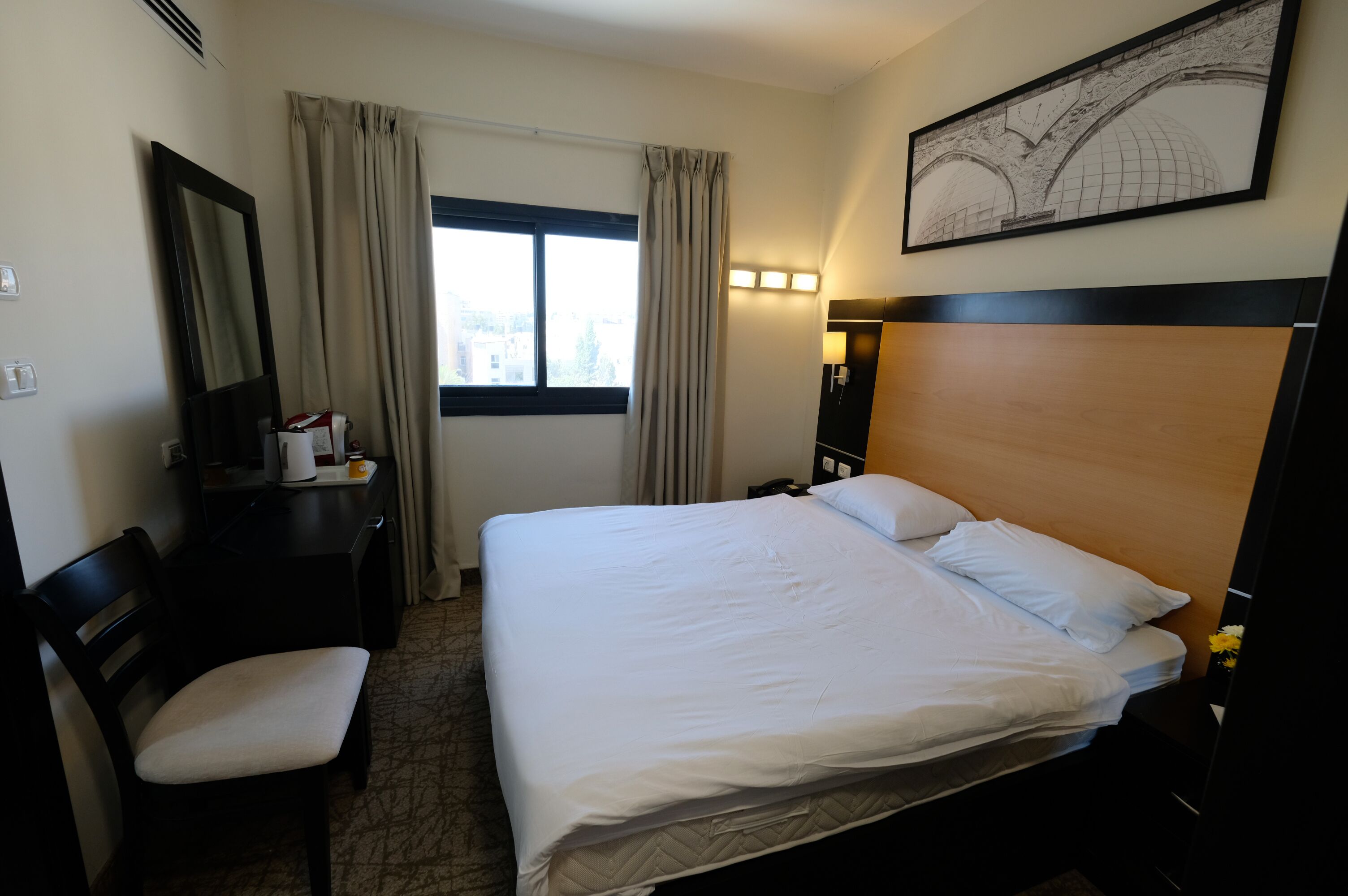 A hotel room with a bed, a desk, a chair, a window with curtains, a wall-mounted lamp, and a framed picture on the wall.