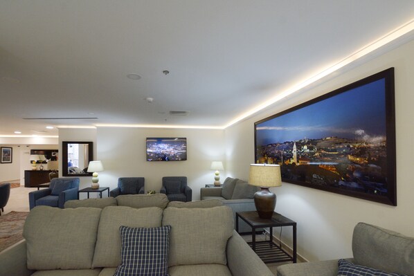 A living room with a large cityscape painting, multiple seating areas, and a view of the city at night. - New Capitol Hotel - Jerusalem (Jerusalem)