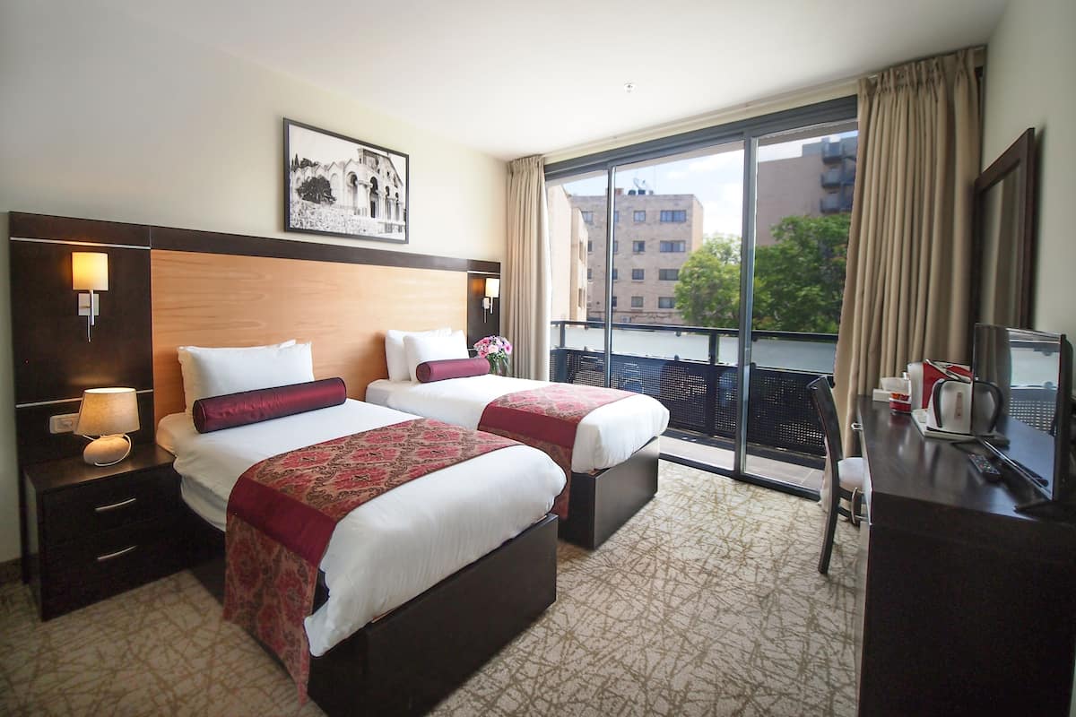 A hotel room with two beds, a desk, a chair, a lamp, a picture on the wall, and a view of the city through the window.