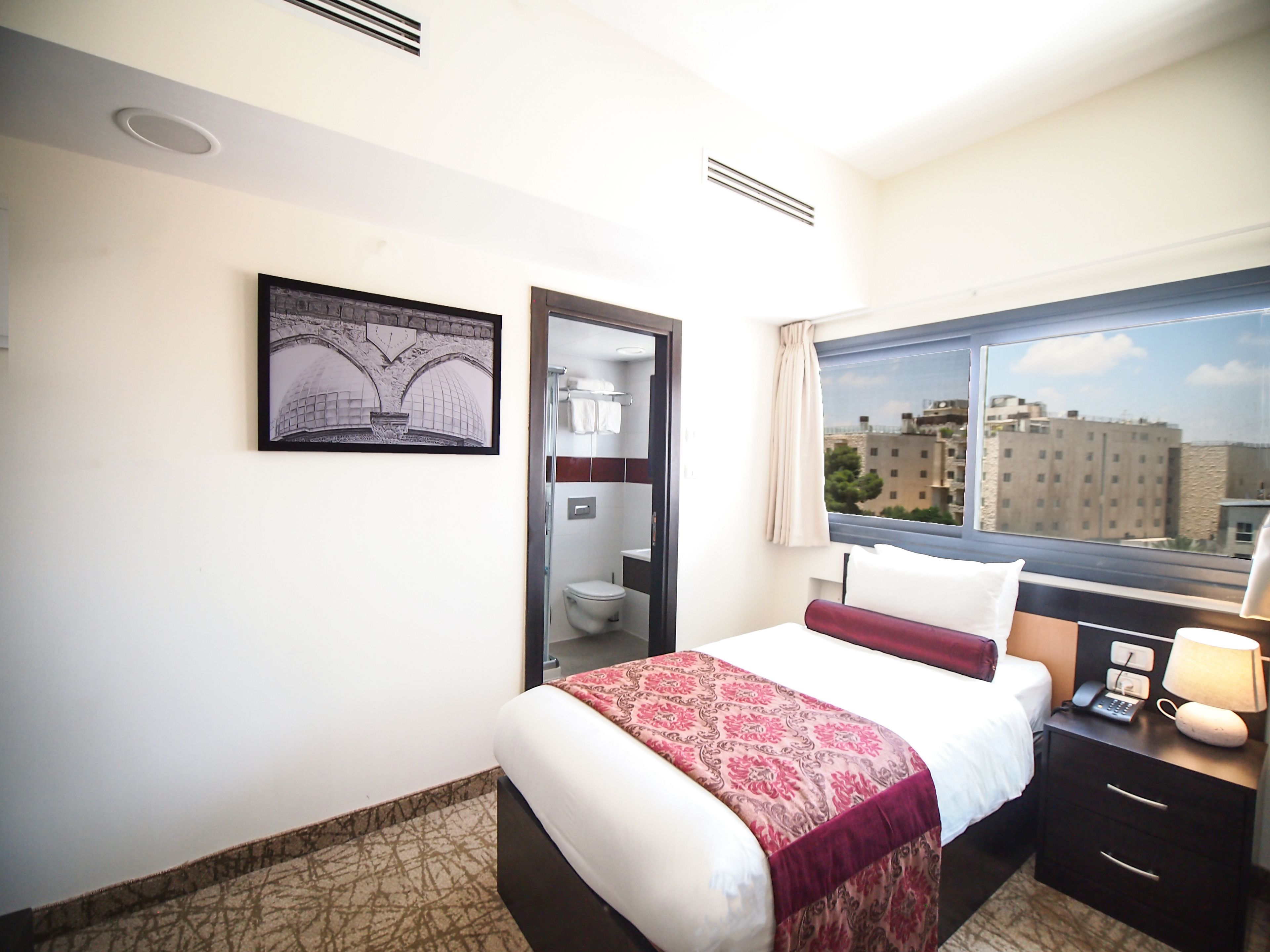 Habitación de hotel con cama, mesita de noche, ventana con vistas a la ciudad, puerta de baño y un cuadro en la pared.