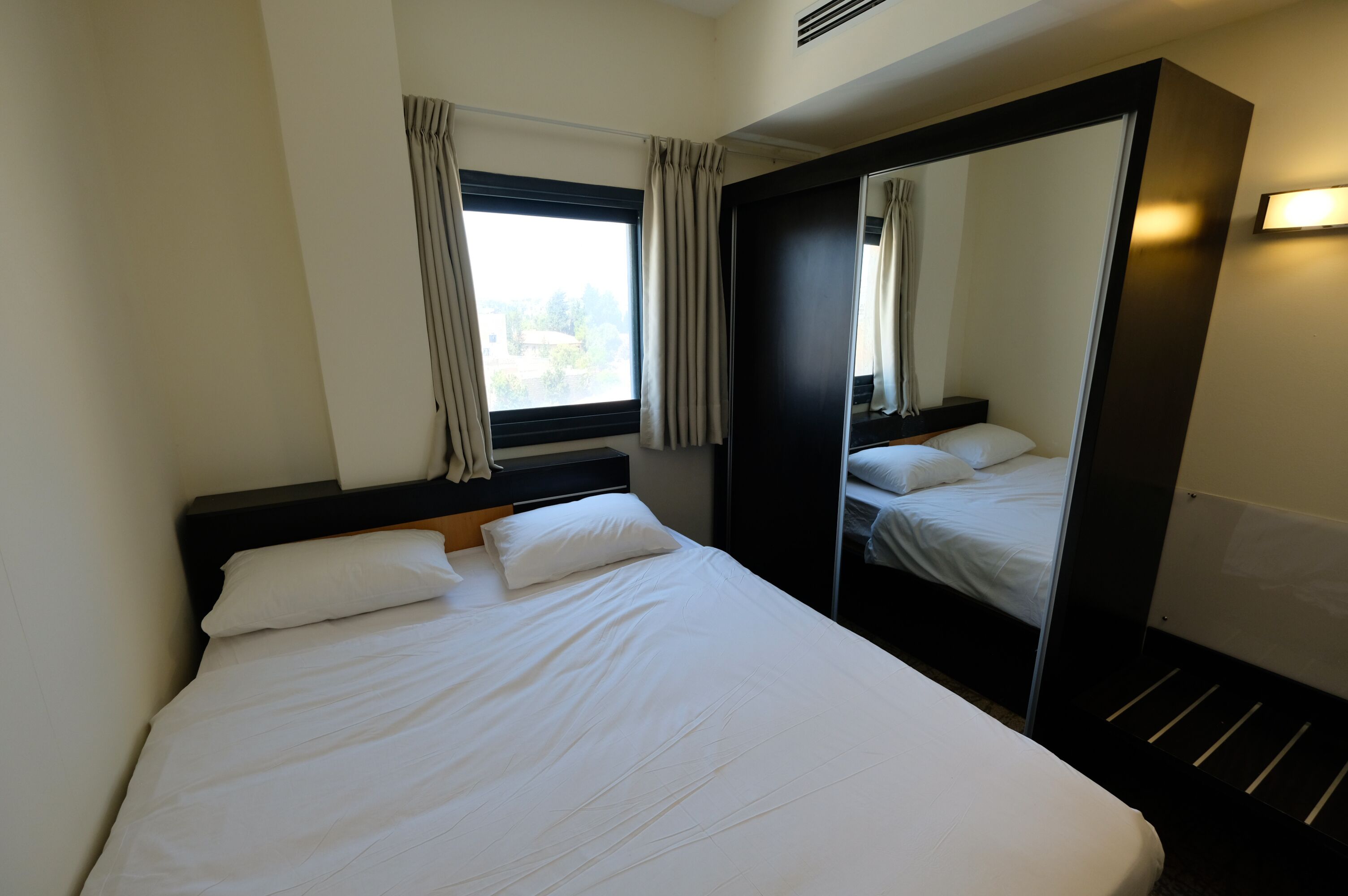 A hotel room with a large bed, a wardrobe, a window with curtains, and a mirror.