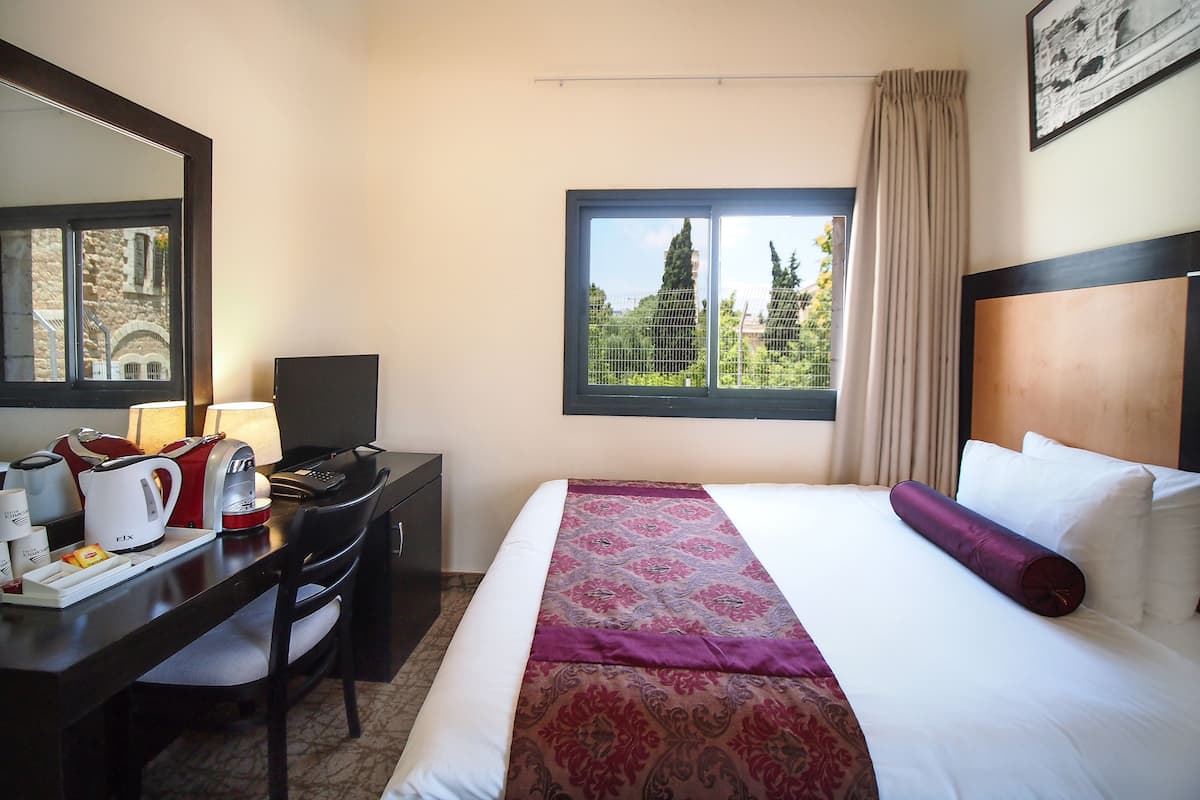 A hotel room with a bed, a desk with a computer, a chair, a lamp, and a window with a view of trees and buildings.