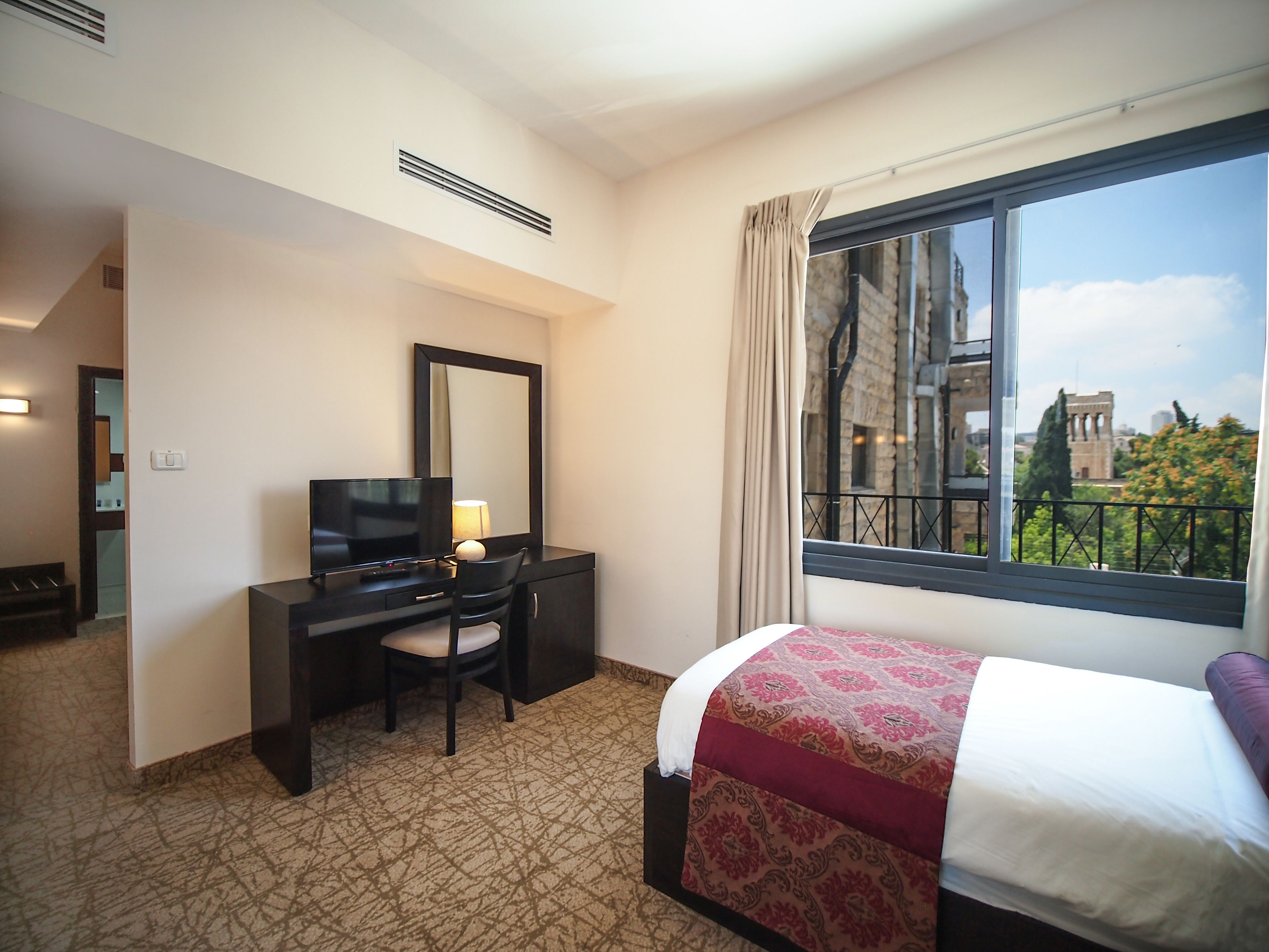 Una camera d'albergo con un letto, una scrivania con un televisore, una sedia, un'ampia finestra vista sulla città e un tappeto decorato.