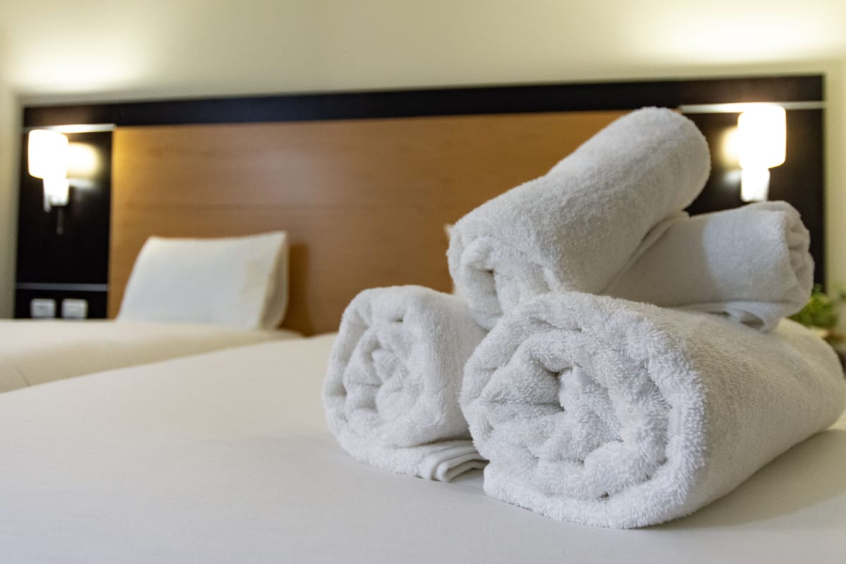 A neatly made bed with white linens and a stack of rolled towels on top.