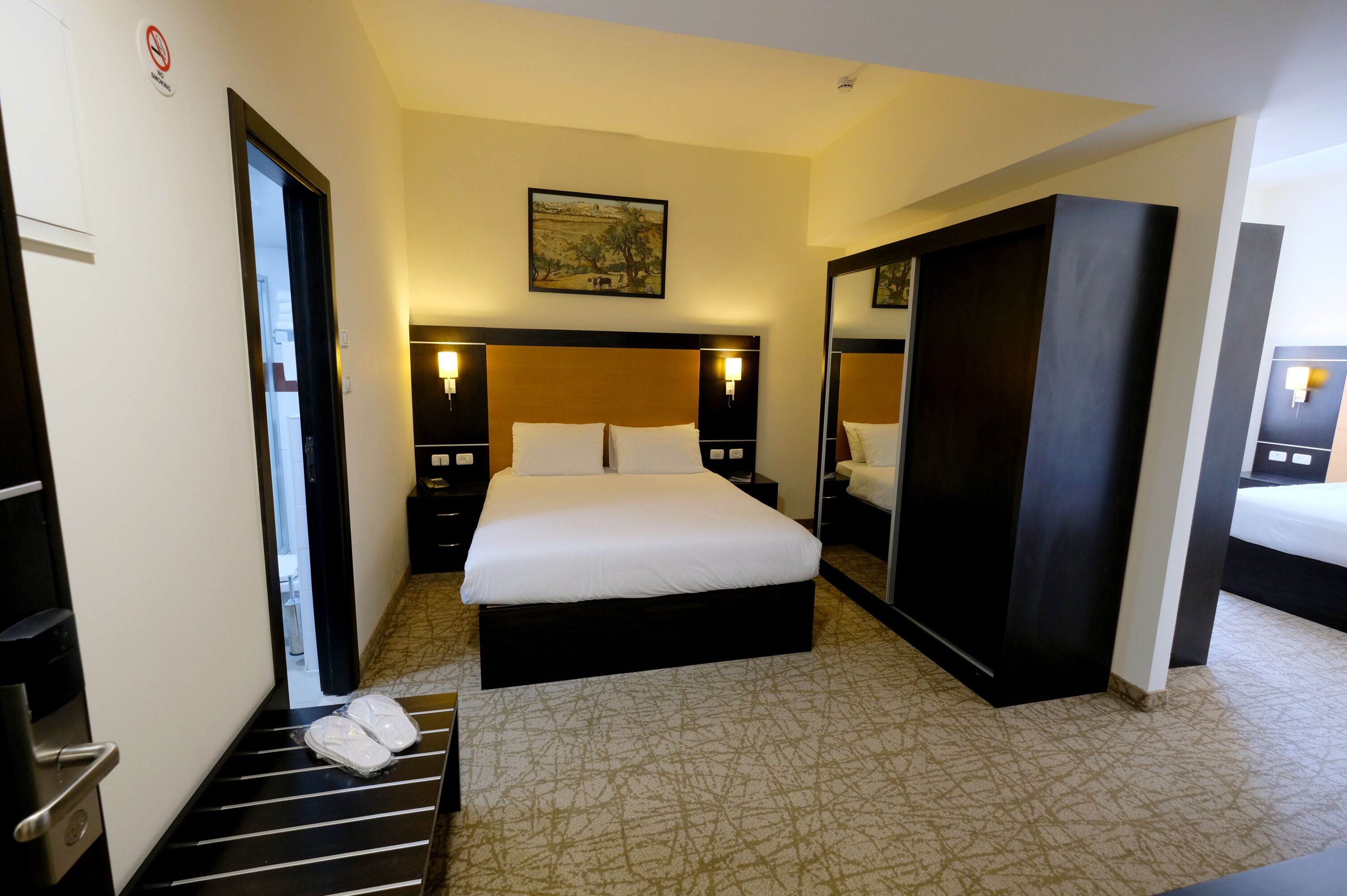 A hotel room with a large bed, two bedside tables, a nightstand, a wardrobe, and a mirror.
