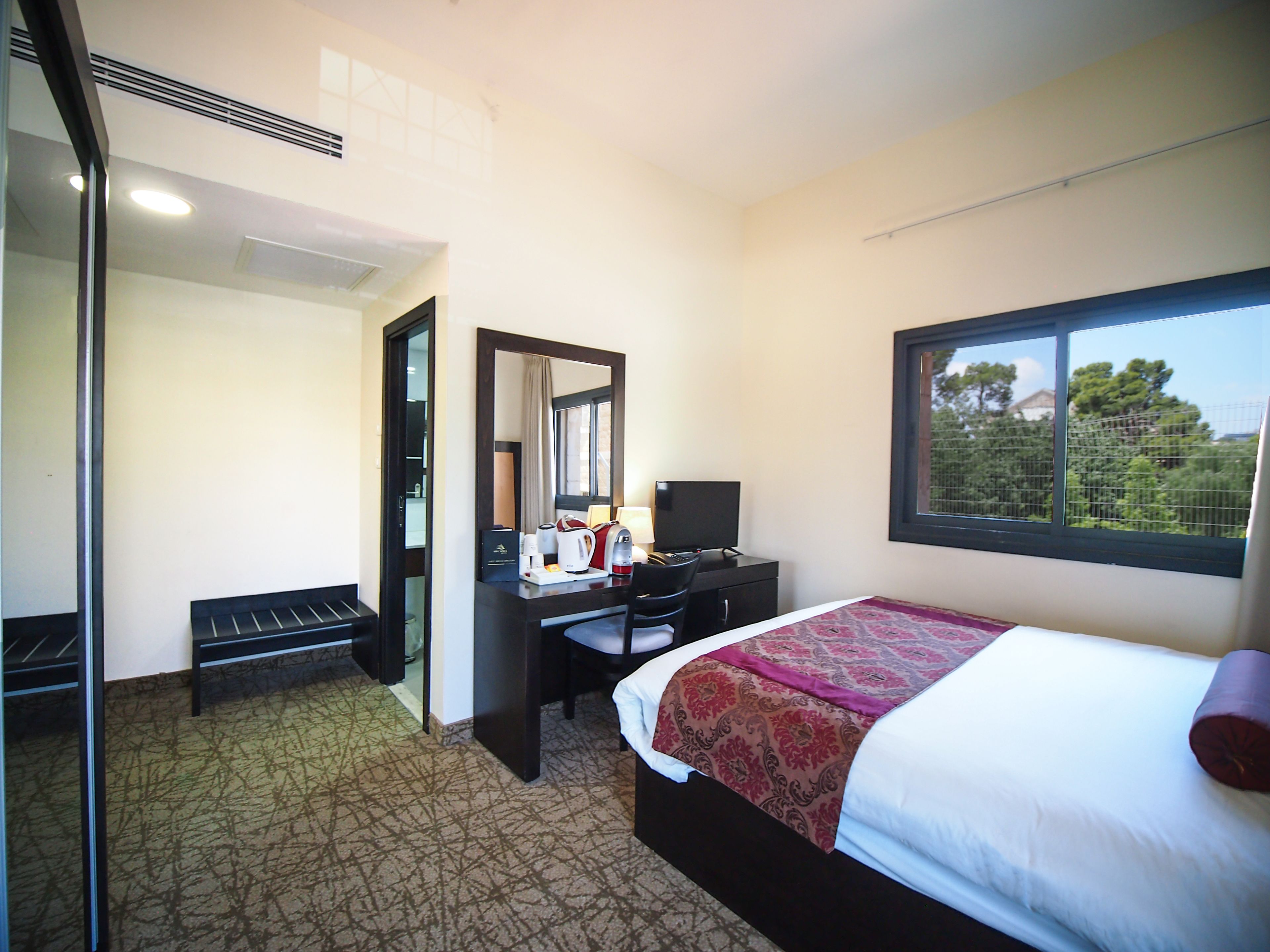 A hotel room with a bed, a desk with a computer, a mirror, and a window with a view of trees.