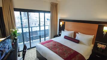 A hotel room with a large bed, a desk with a computer, and a view of the city through the windows.