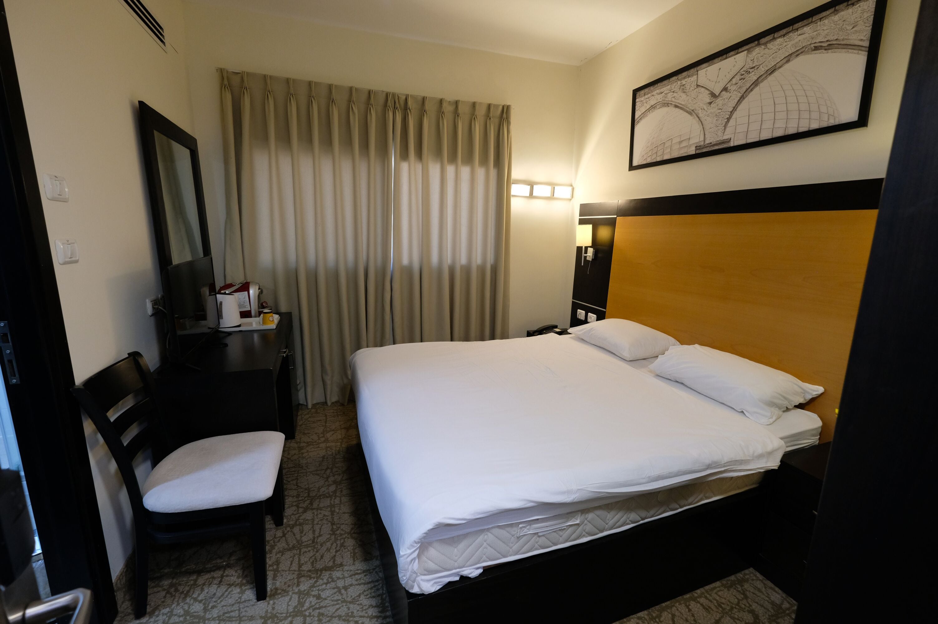 Quarto de hotel com uma cama, uma escrivaninha com cadeira, um espelho, um abajur e uma janela com cortinas.