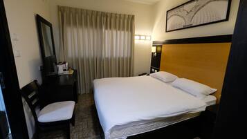 Quarto de hotel com uma cama, uma escrivaninha com cadeira, um espelho, um abajur e uma janela com cortinas.