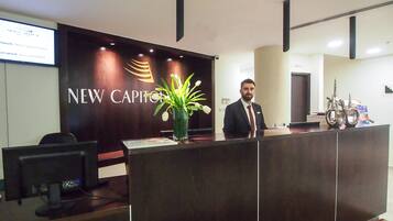 A reception desk with a 'New Capitol' sign, a man standing behind it, and a laptop on the desk.