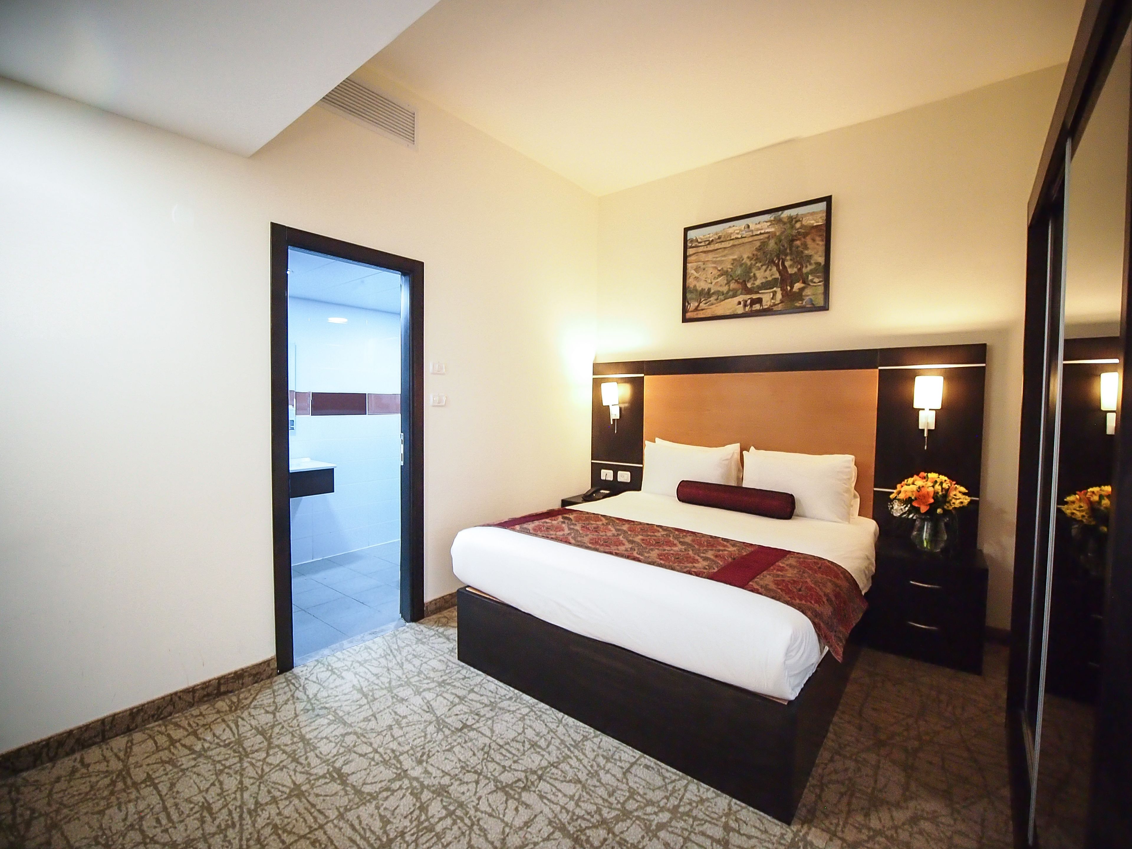 Une chambre d’hôtel avec un grand lit, une tête de lit agrémentée de lampes murales, un tableau au-dessus du lit, une porte donnant sur une autre pièce et une petite table sur laquelle est posé un vase de fleurs.