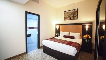 Une chambre d’hôtel avec un grand lit, une tête de lit agrémentée de lampes murales, un tableau au-dessus du lit, une porte donnant sur une autre pièce et une petite table sur laquelle est posé un vase de fleurs.