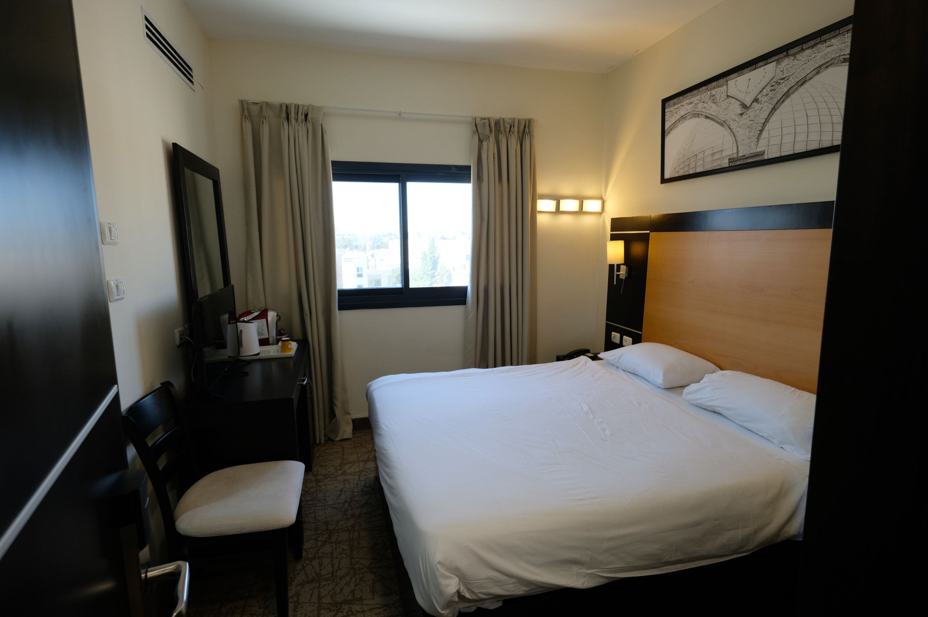 Hotelkamer met een bed, bureau, stoel, raam met gordijnen, een wandlamp en een lijst met een afbeelding.