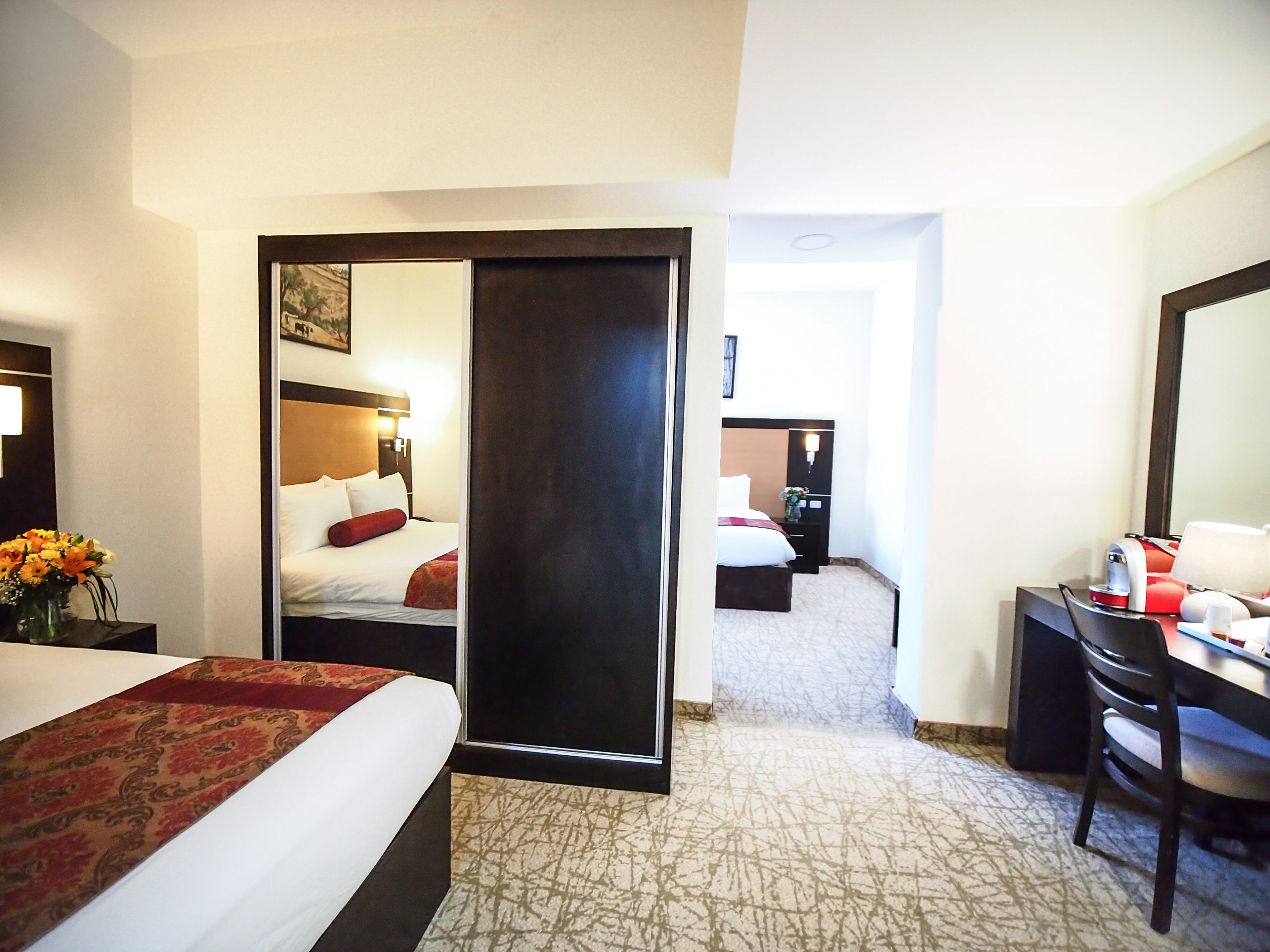 Ein Hotelzimmer mit einem Bett, einem Schreibtisch und einem Stuhl. An der Wand hängt ein Spiegel und eine Tür führt in einen weiteren Raum.