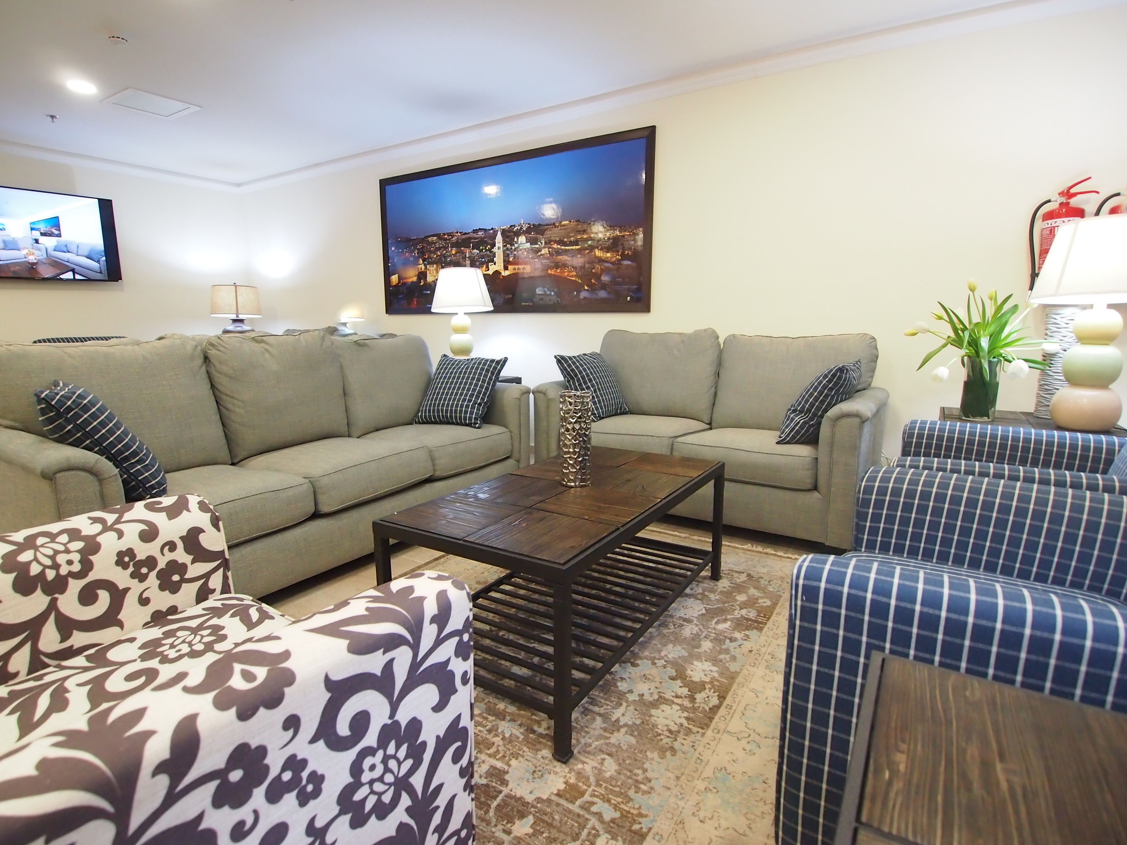 A living room with two sofas, a coffee table, patterned armchairs, a rug, and a cityscape painting on the wall.