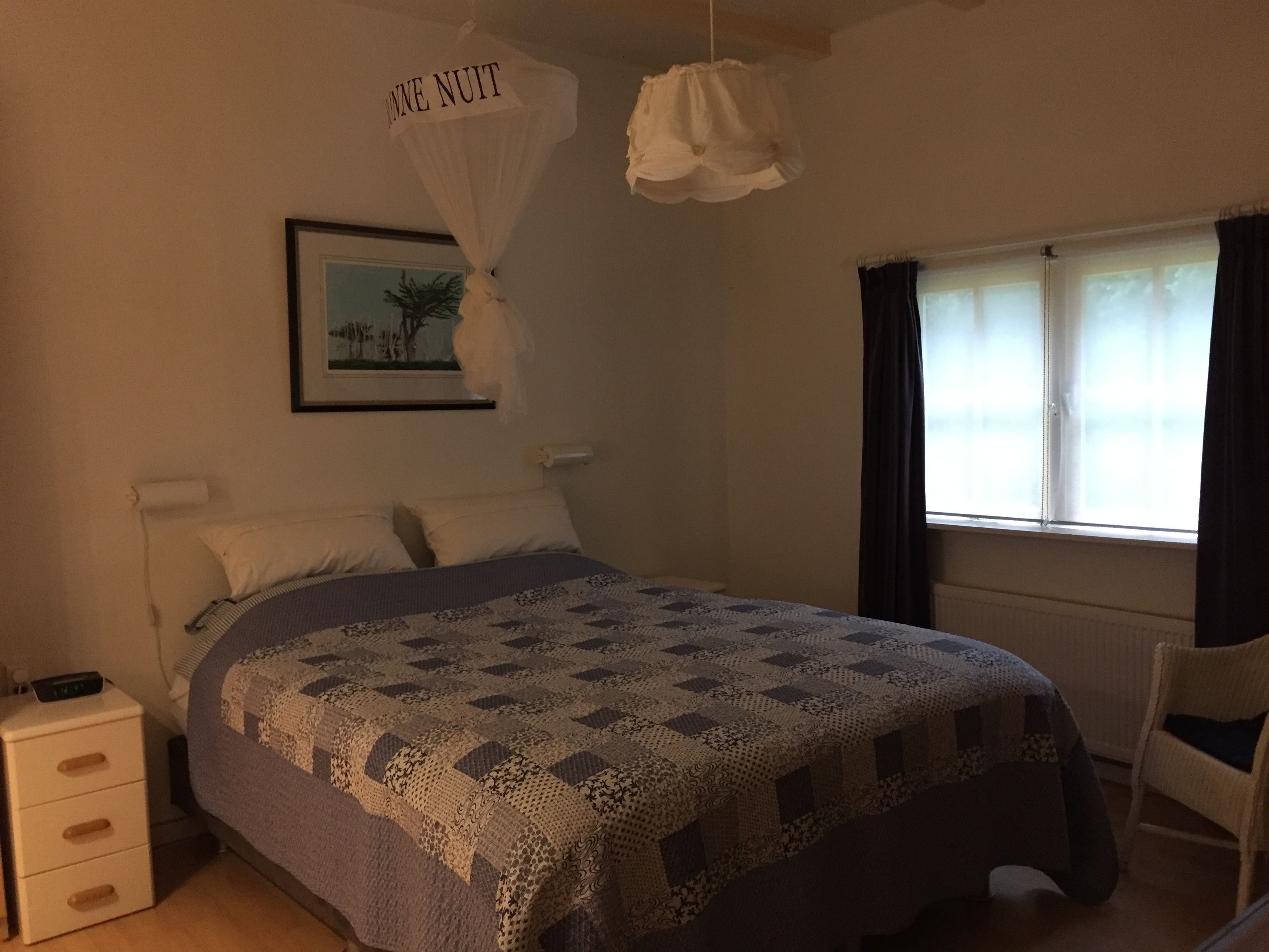 A bedroom with a bed, a bedside table, a chair, a window with curtains, a hanging light fixture, and a wall-mounted picture.