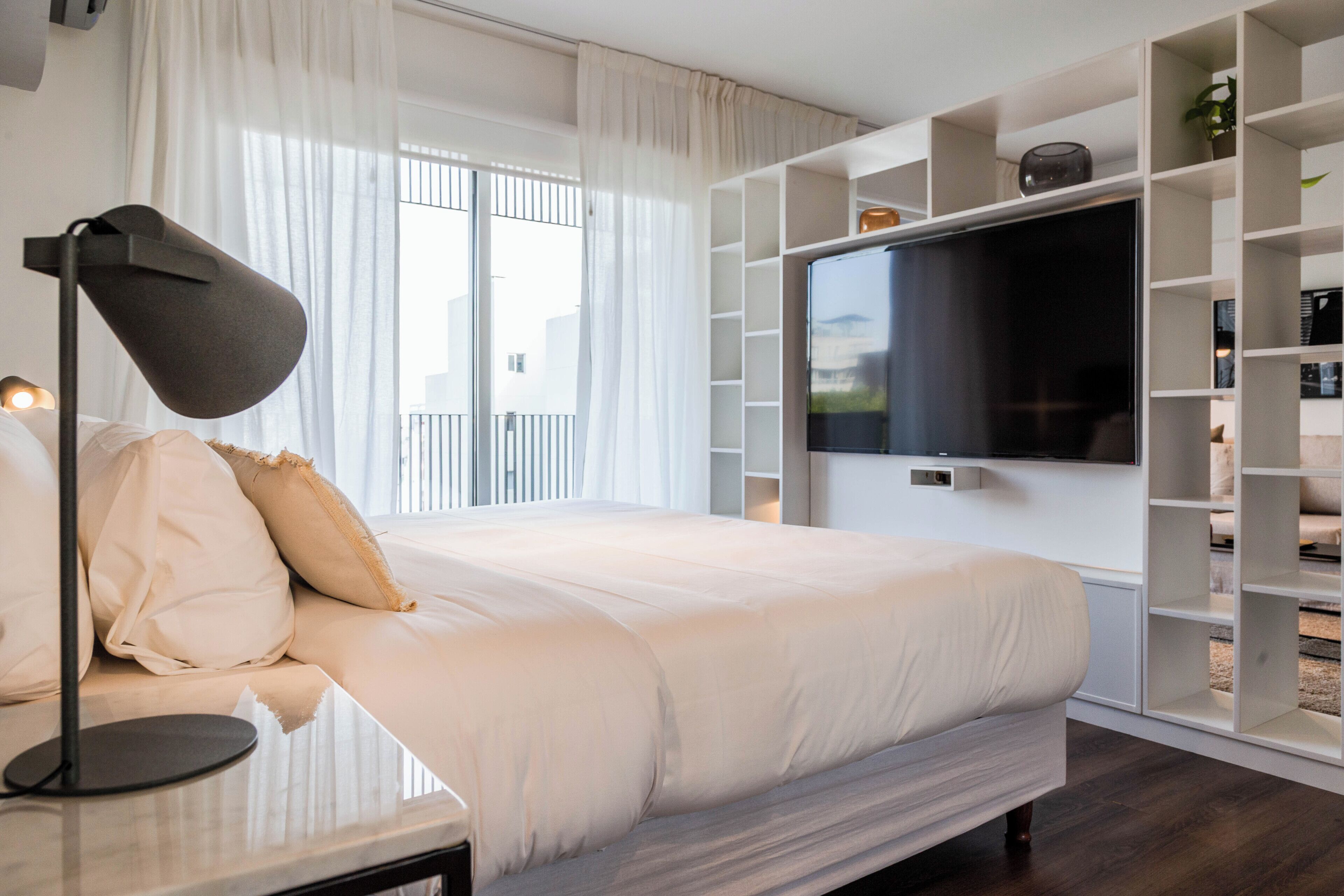 A modern bedroom with a large bed, a flat-screen TV mounted on a white wall, and a built-in bookshelf.