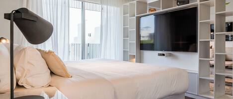 A modern bedroom with a large bed, a flat-screen TV mounted on a white wall, and a built-in bookshelf.